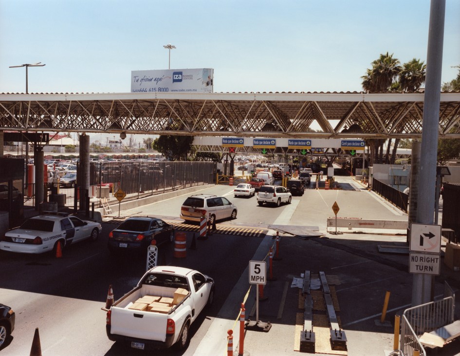 Vehicles queuing at a US-Mexico border checkpoint with metal barriers and security infrastructure.