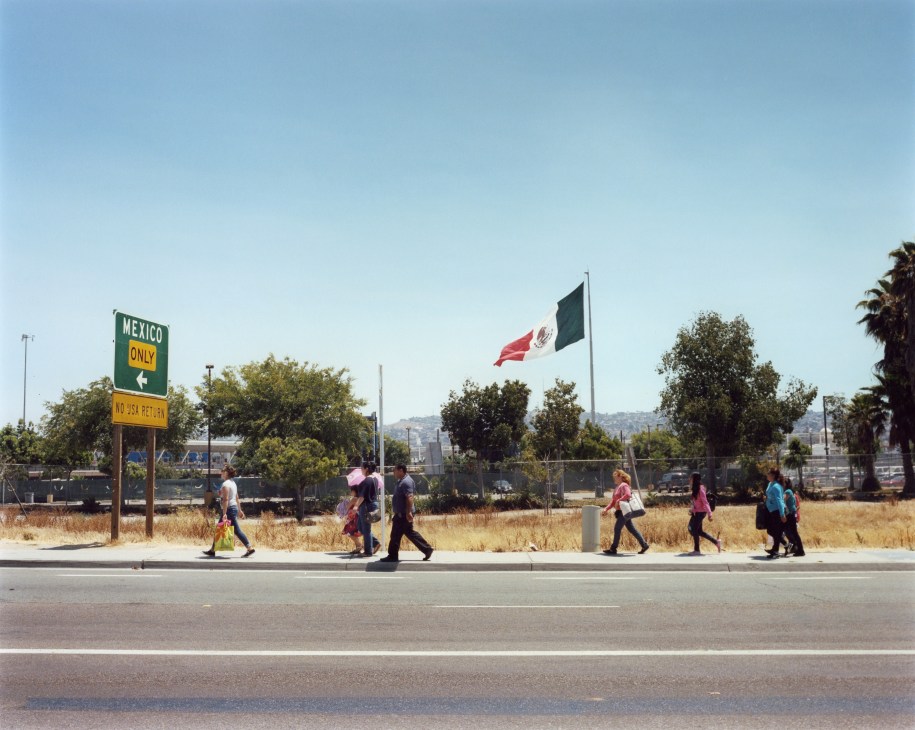 Pedestrians cross at the US-Mexican border with Mexico-only sign and Mexican flag visible.
