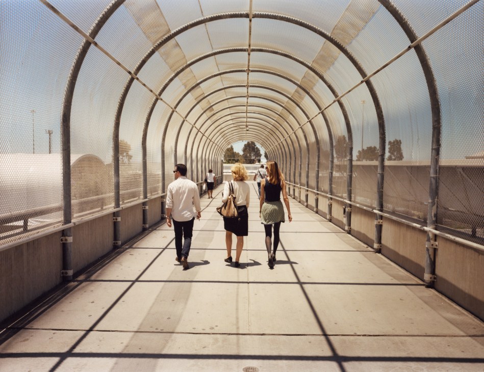 Pedestrians walking through a covered bridge or tunnel with arched metal framework on the US side of the border.