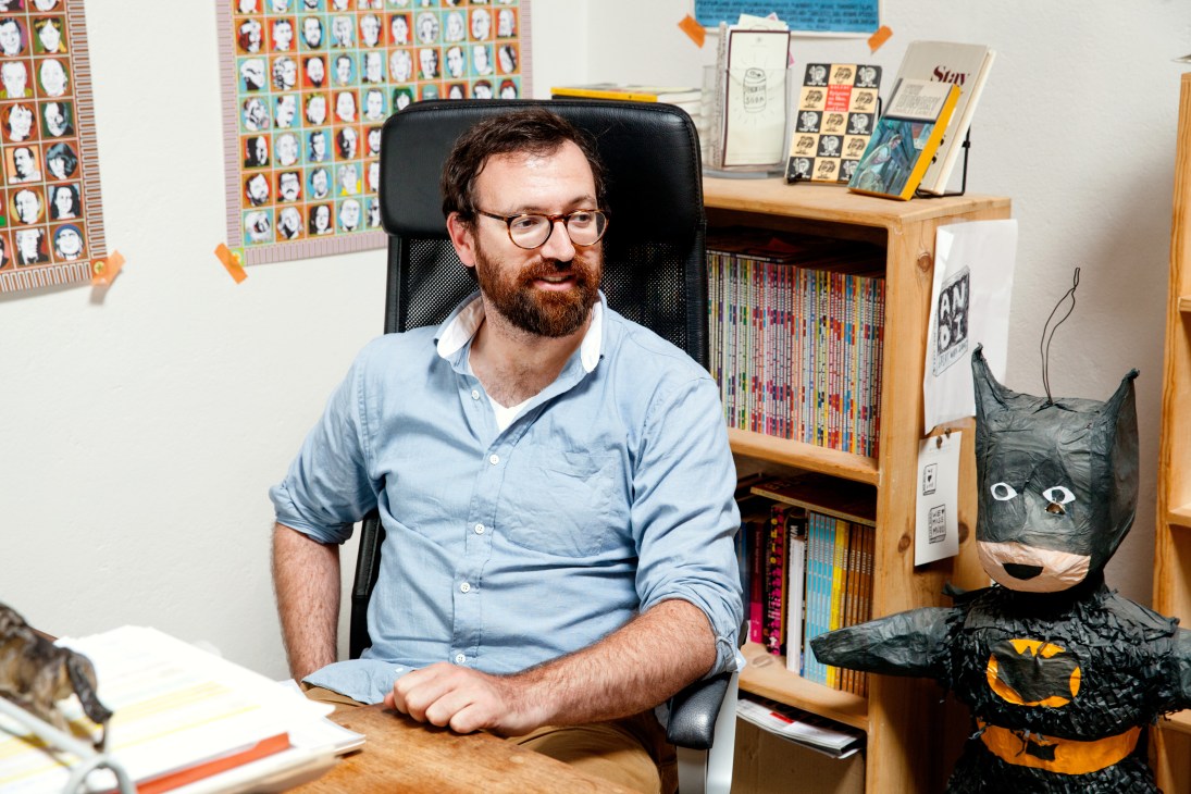 Man in light blue shirt seated at desk in office with bookshelves and artwork behind him.