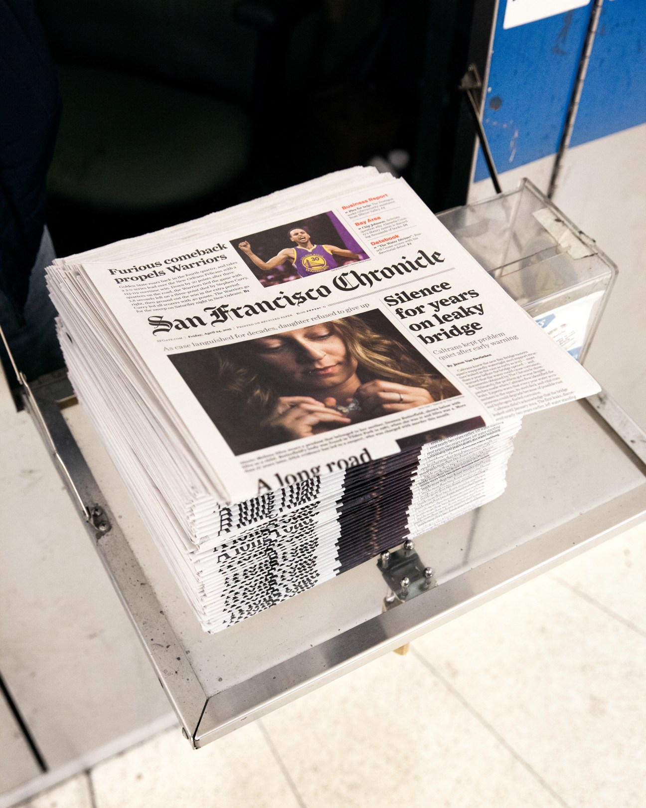 Stacked newspapers at Montgomery Street Station newsstand displaying San Francisco Chronicle front pages with various headlines and images.