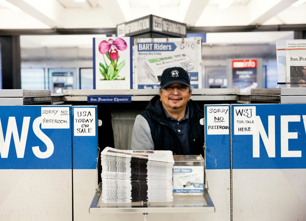 Newsstand vendor at Montgomery Street Station with stacked newspapers displayed in front of blue signage