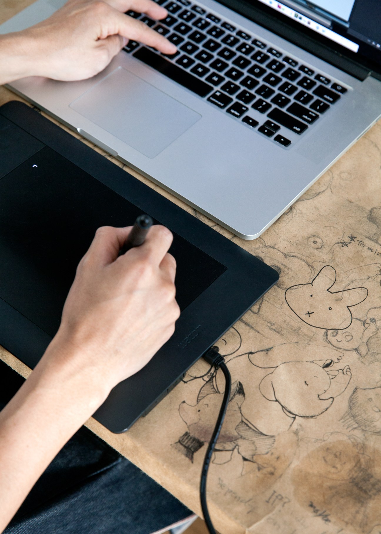 Person working at desk with laptop and drawing tablet, surrounded by sketched character drawings on kraft paper.