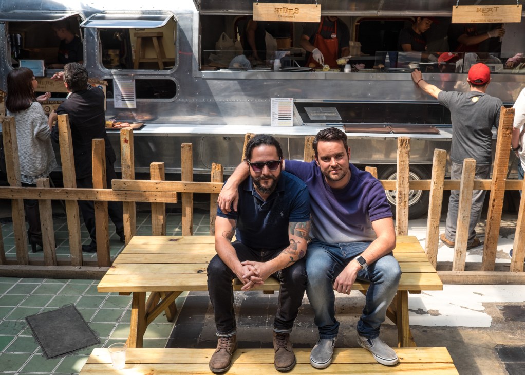 Two men sit on a wooden bench in front of Pinche Gringo BBQ's food trailer in Mexico City.