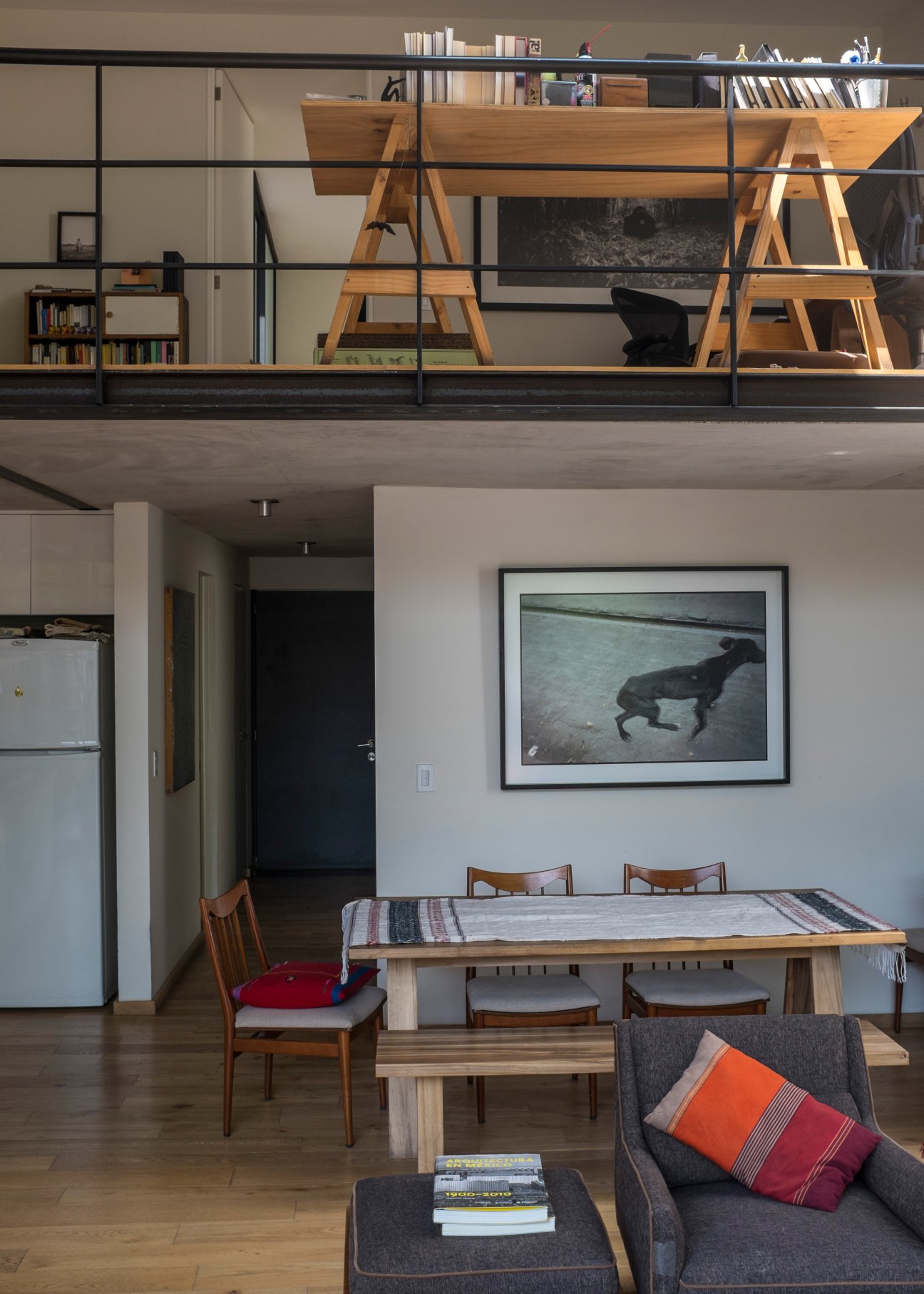 Modern loft interior with mezzanine level, wooden shelving, dining area with mid-century chairs, and framed artwork on white walls.