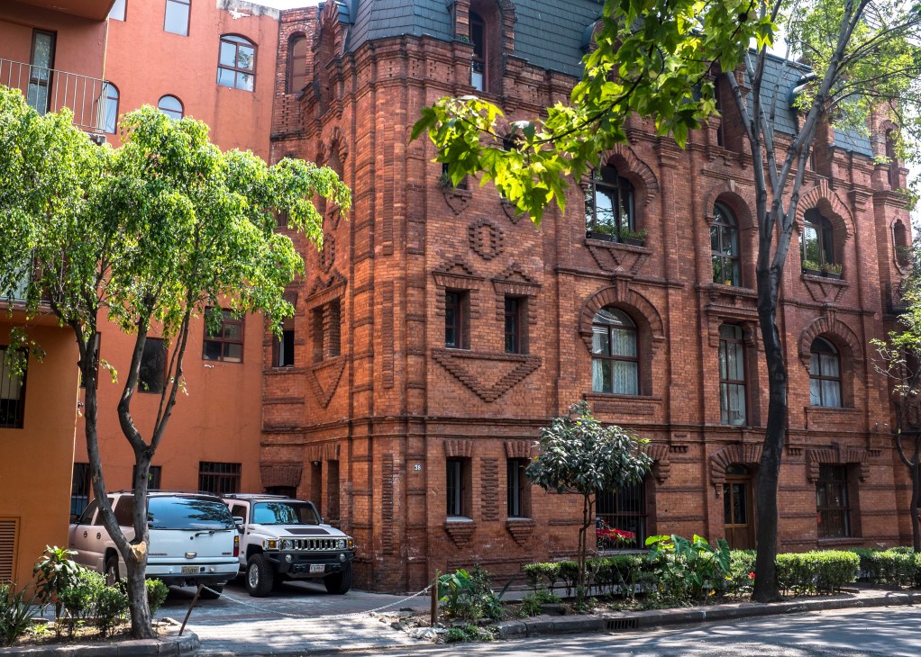 Red brick building with arched windows and decorative detailing, surrounded by trees and parked cars in Mexico City's Juárez neighbourhood.