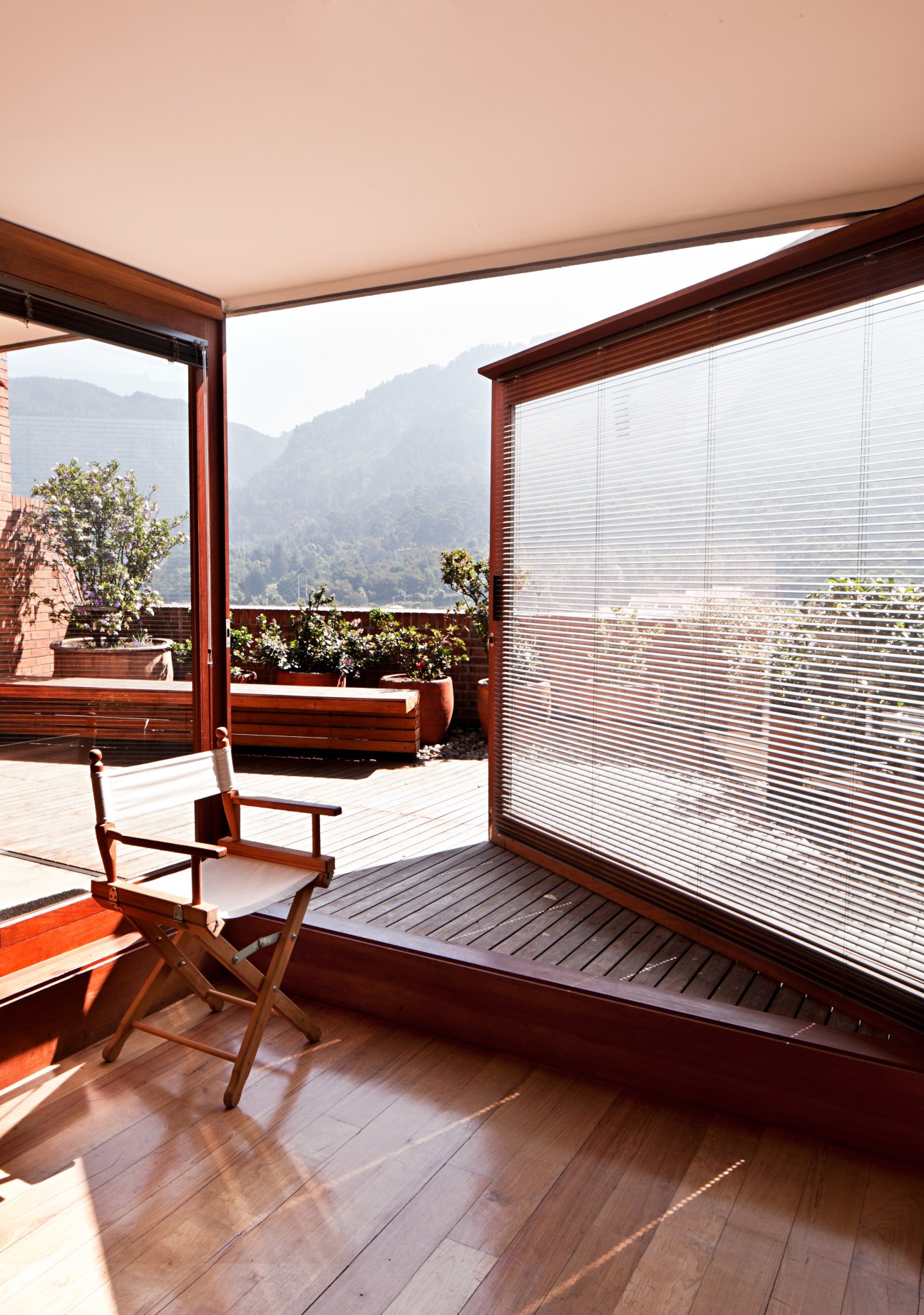 A wooden terrace with mountain views, featuring a chair and potted plants with louvered screens.