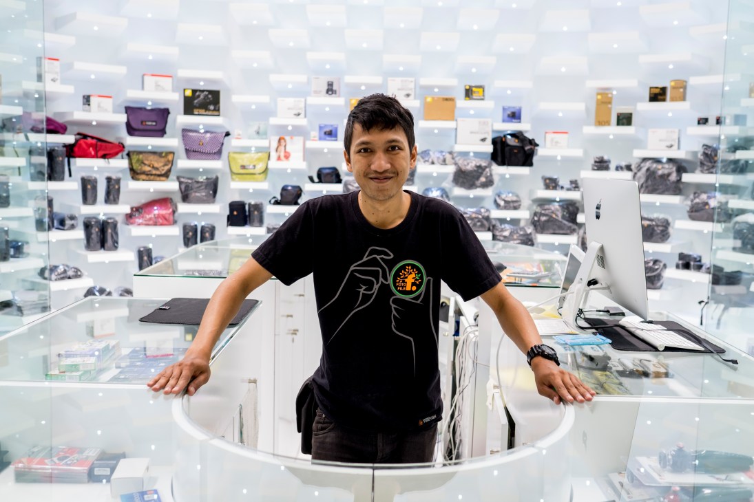 Manager at Foto File store in Central Embassy with colorful handbags and merchandise displayed on white shelving behind him.