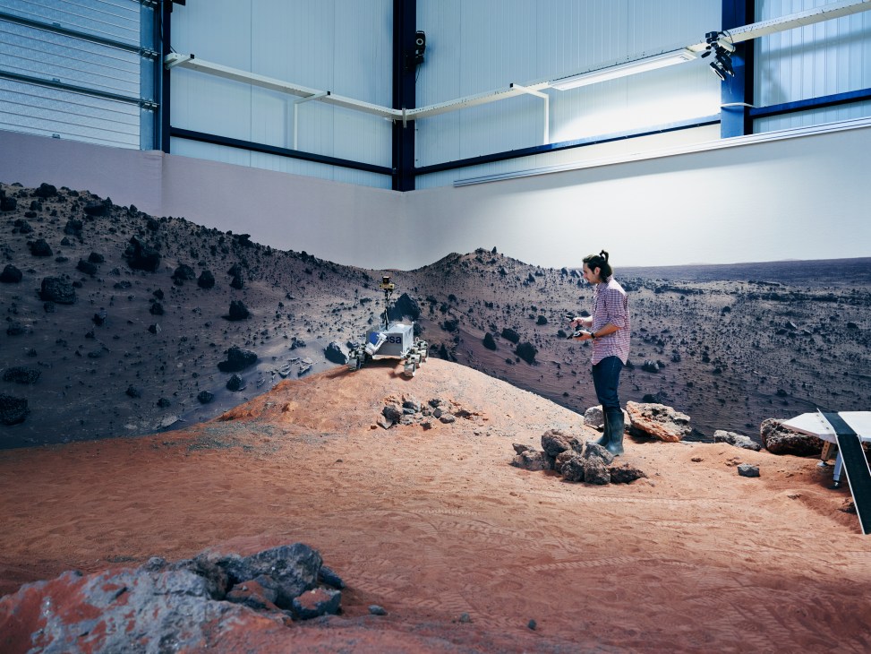 Engineer operates a rover in a red dust-covered Mars simulation yard inside a large hangar facility.
