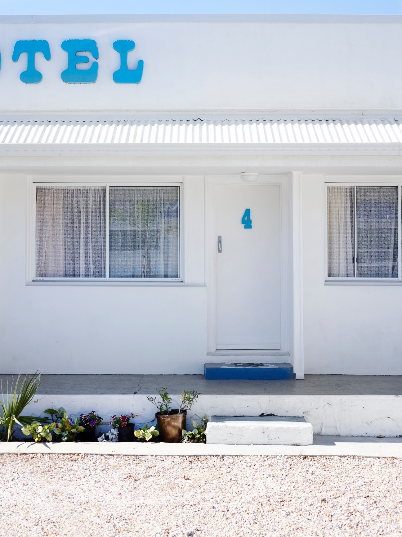 White mid-century motel facade with blue signage and number 4, featuring windows with patterned curtains and flowering planters.