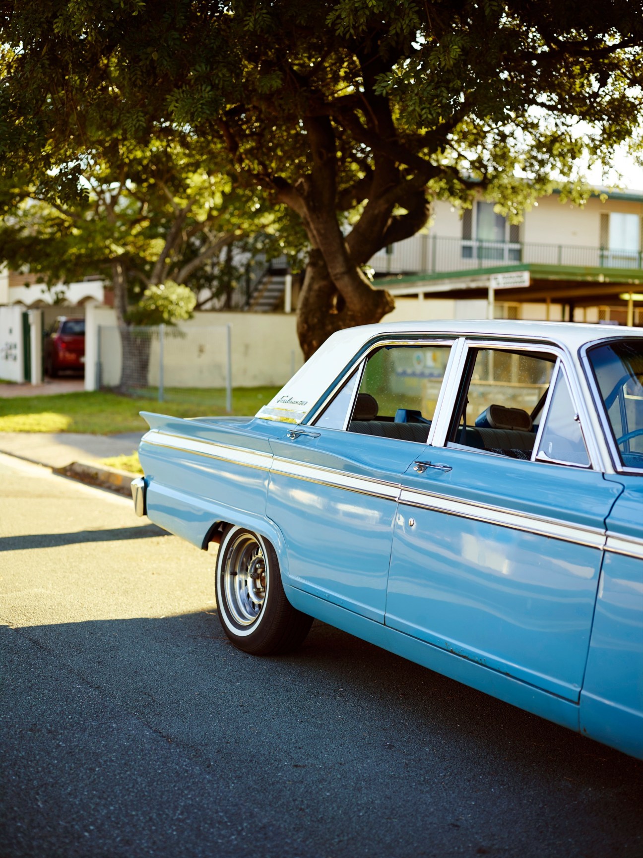 Light blue classic 1960s Ford Fairlane parked on a driveway beneath a large tree, with mid-century homes visible in the background.
