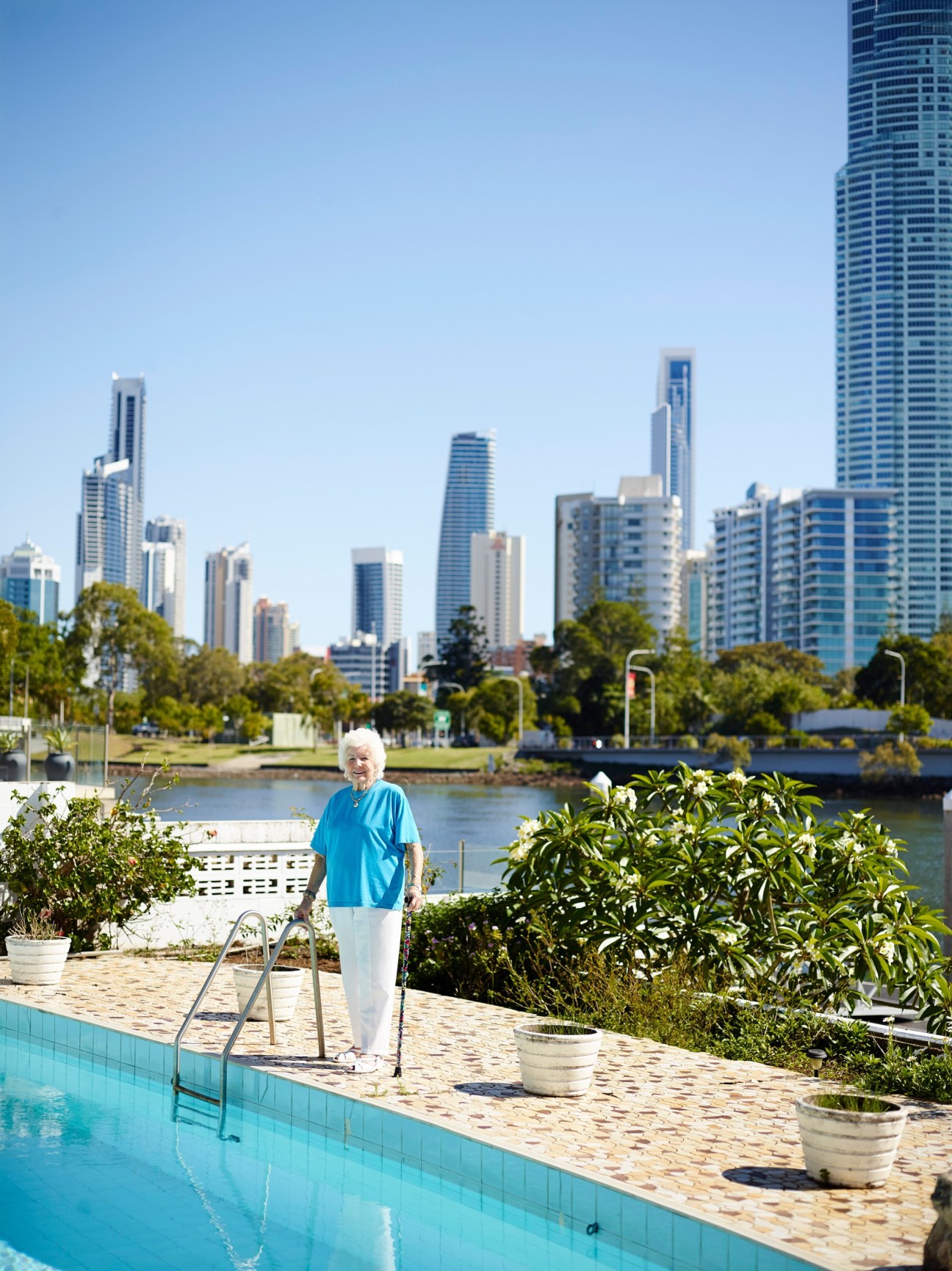 Elderly woman in blue shirt standing by a pool overlooking a waterway with high-rise buildings in the background.