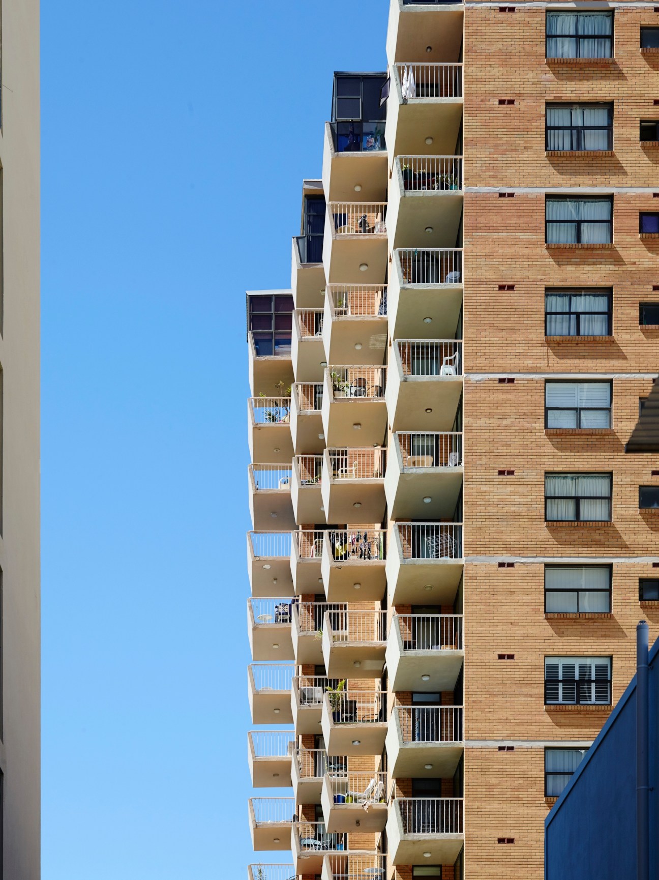 Multi-story apartment building with red brick facade and protruding balconies against clear blue sky.