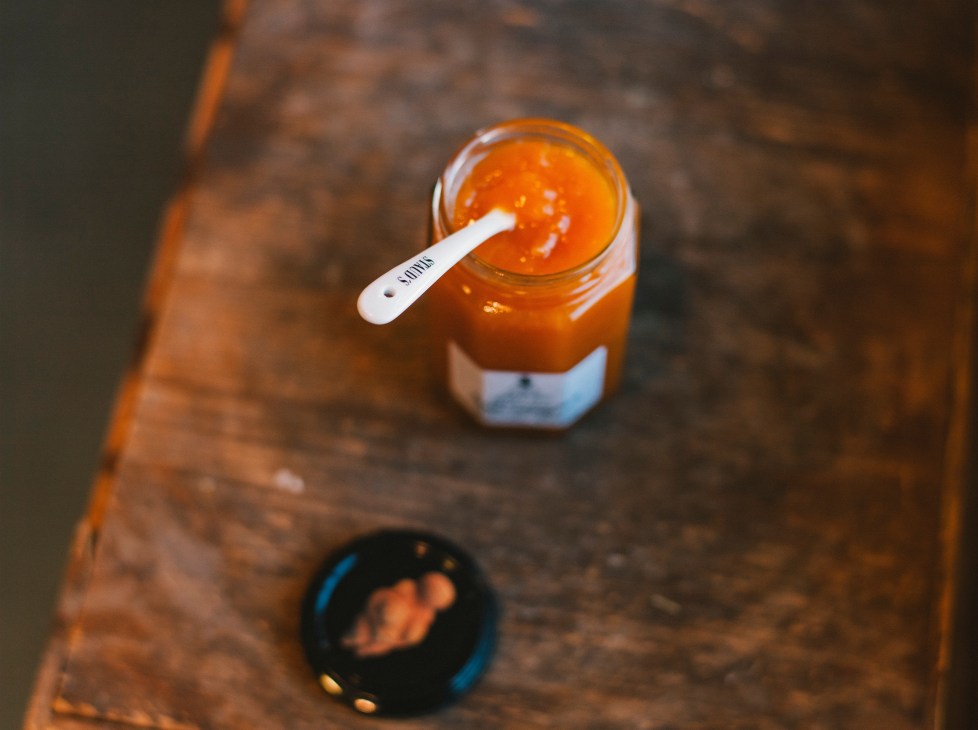 Jar of orange Staud's preserve with white spoon on rustic wooden surface.