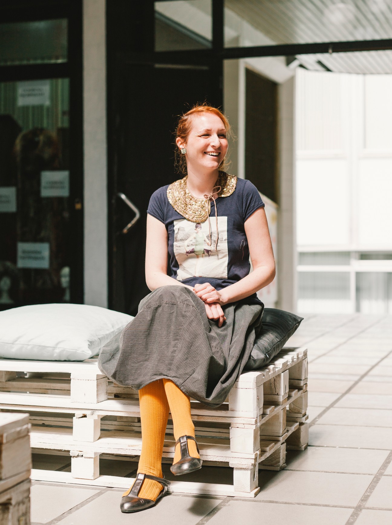 Fashion designer Aleksandra Lalic sits smiling on a white wooden pallet bench in a bright, minimalist space.