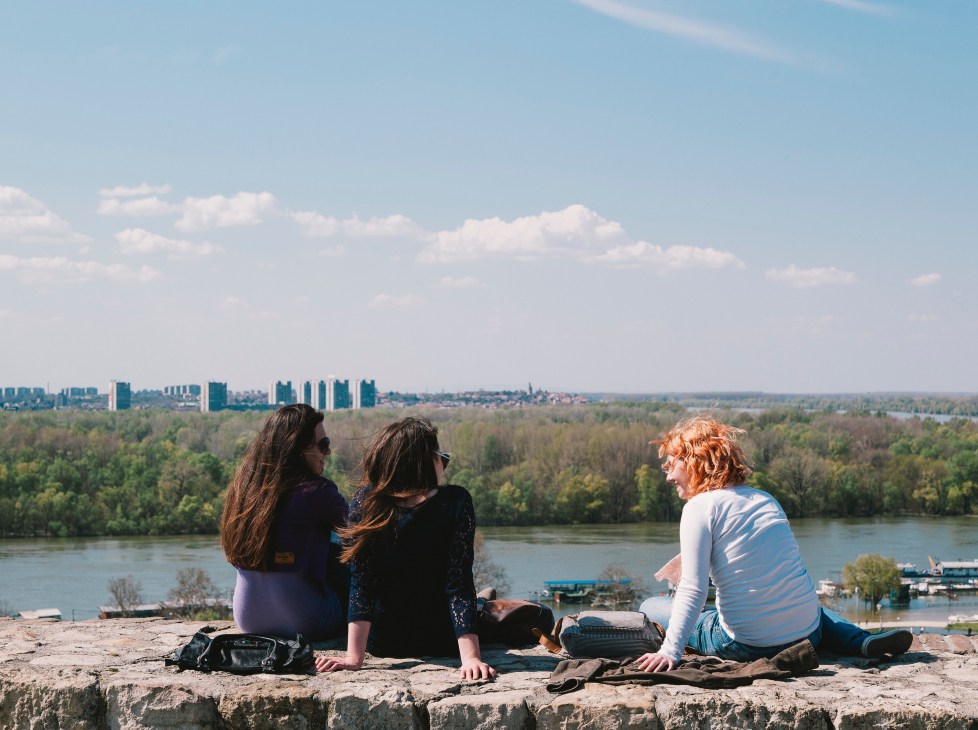 Three women sitting on a stone ledge overlooking the Sava river, Novi Beograd skyline and green forests.