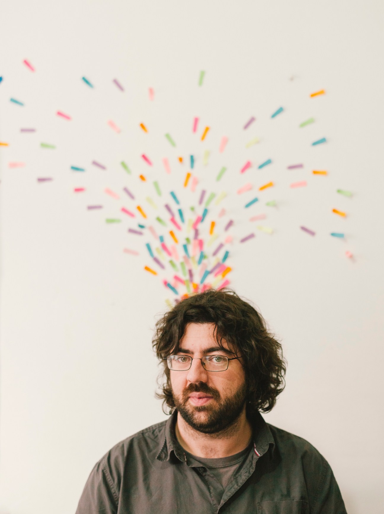 Man with long dark hair and glasses wears a dark shirt against a white wall with colorful confetti-like streaks radiating from his head.