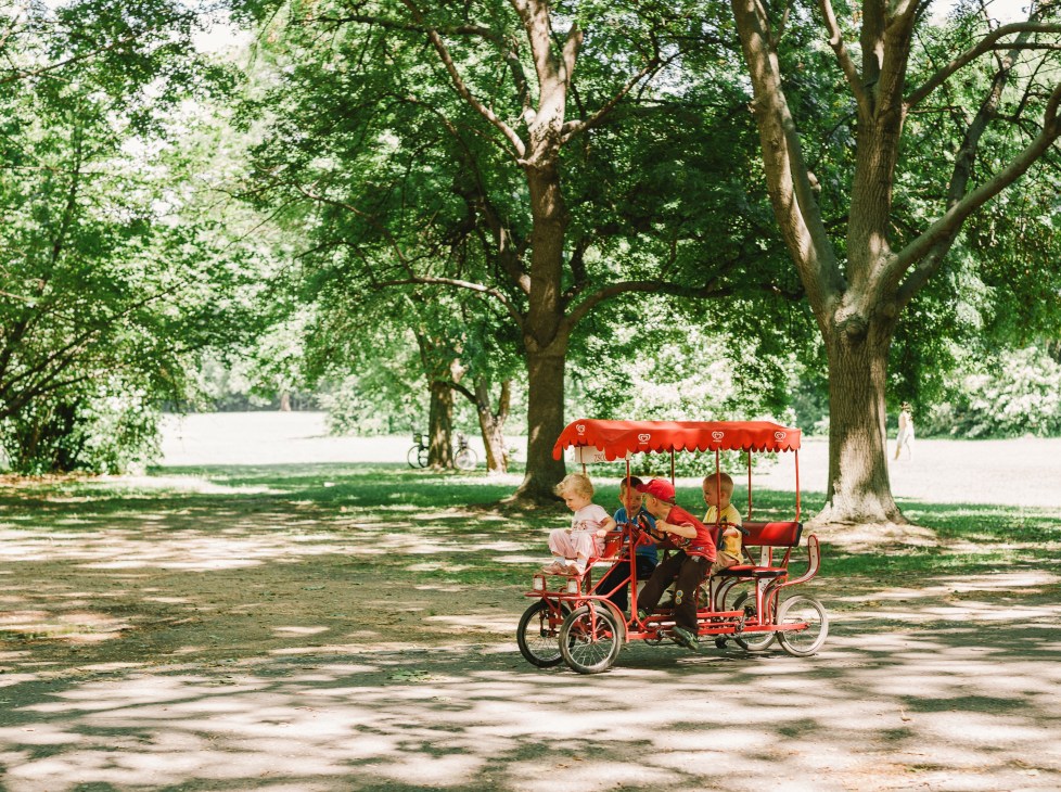 A red pedalo bike with canopy carries four children under large shade trees on Margaret Island.