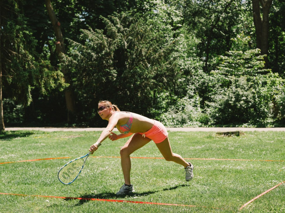Woman playing tennis on a grass court at Margaret Island, Budapest, with trees in the background.