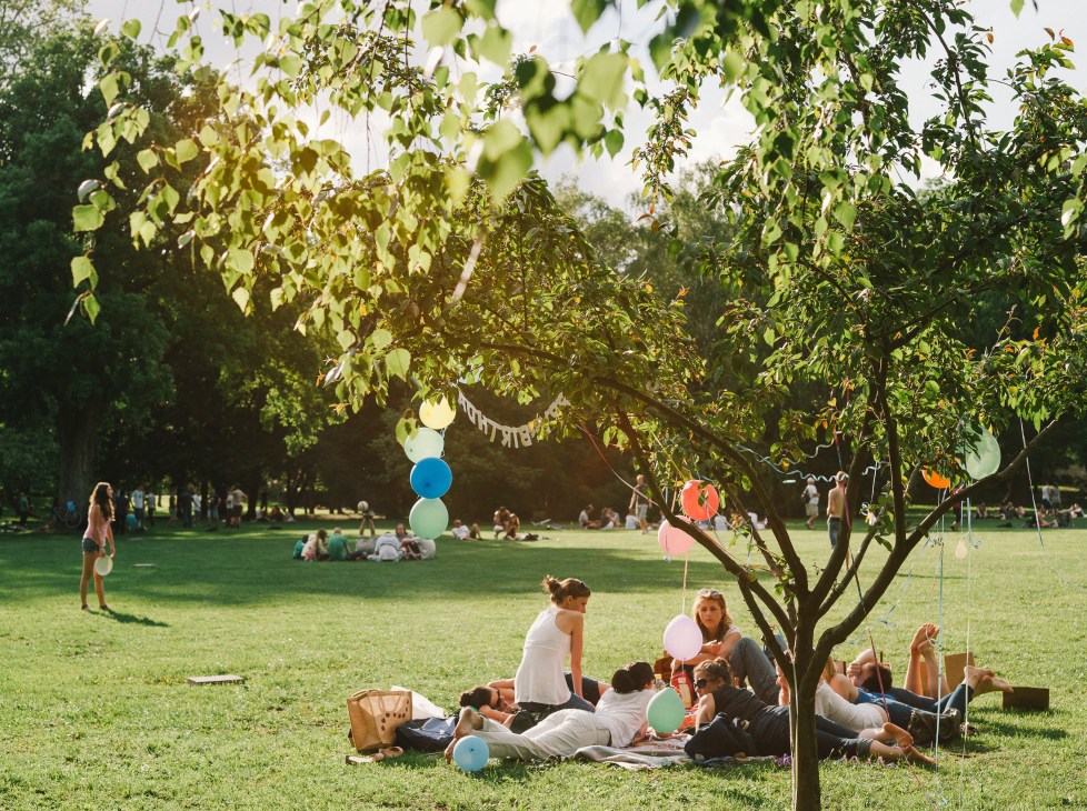 Families and groups picnic under trees on Margaret Island with colorful balloons hanging from branches.