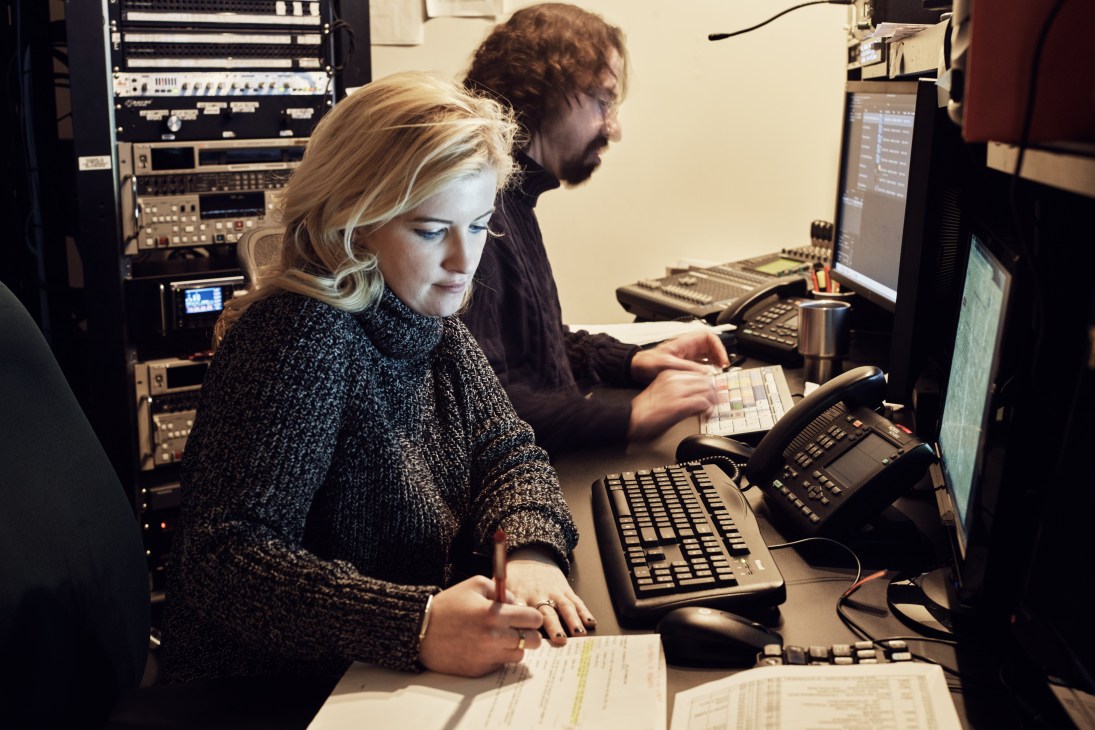 Two people working at computer terminals in a broadcast control room with audio equipment visible.