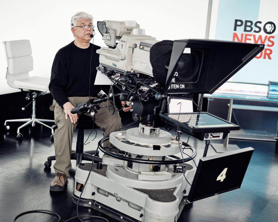 A man in headphones operates a Sony television camera in a PBS Newshour studio control room.