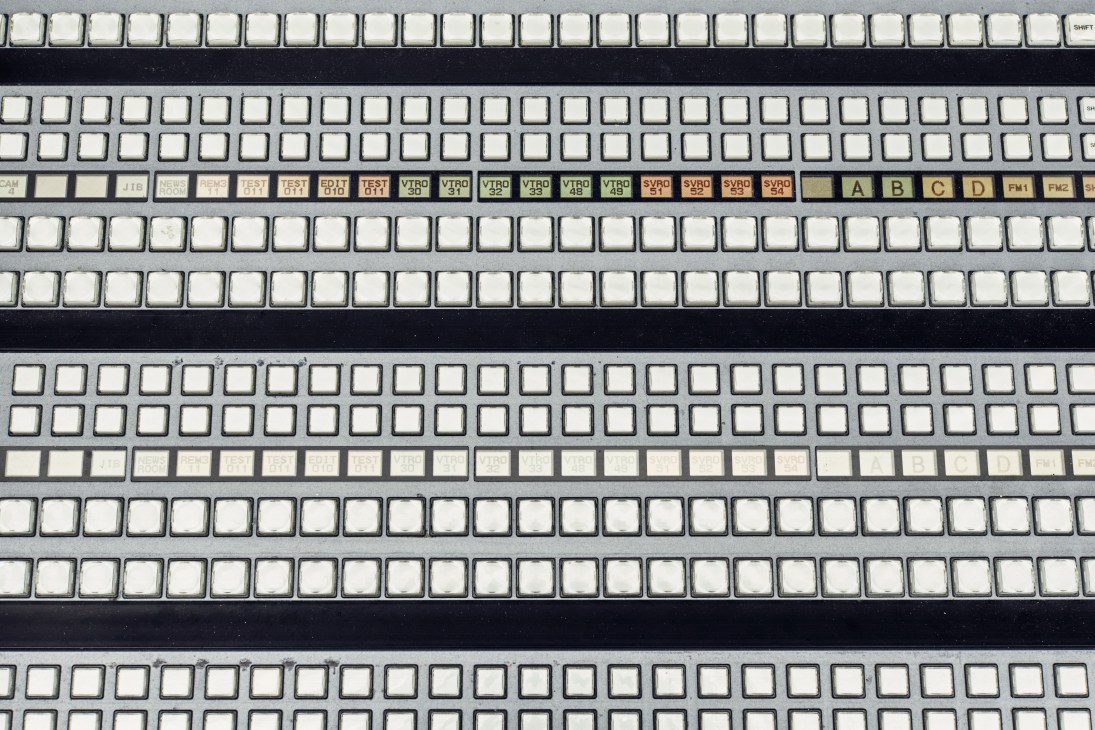 Operating desk in a television control room with multiple rows of white buttons and labeled function keys.