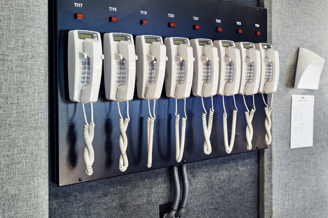 Row of eight white telephone handsets with coiled cords mounted on a blue panel with red indicator lights and numbered labels above.