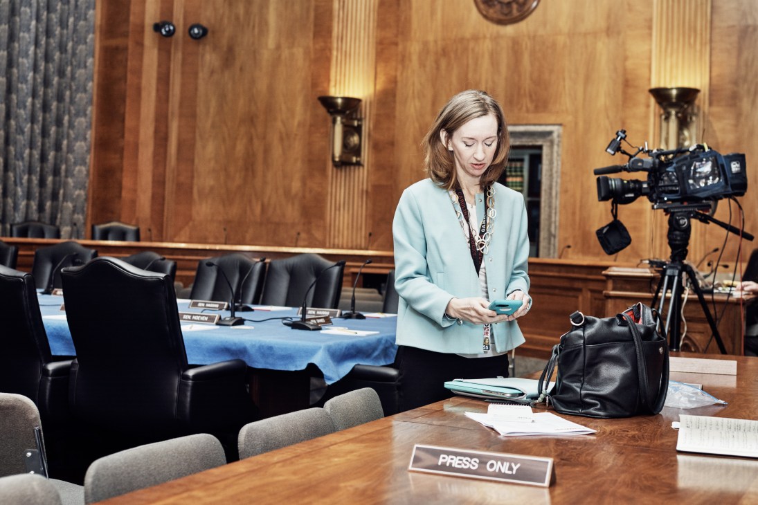A woman in a light blue blazer stands in a Senate committee room holding a phone, with a video camera and press credentials visible.