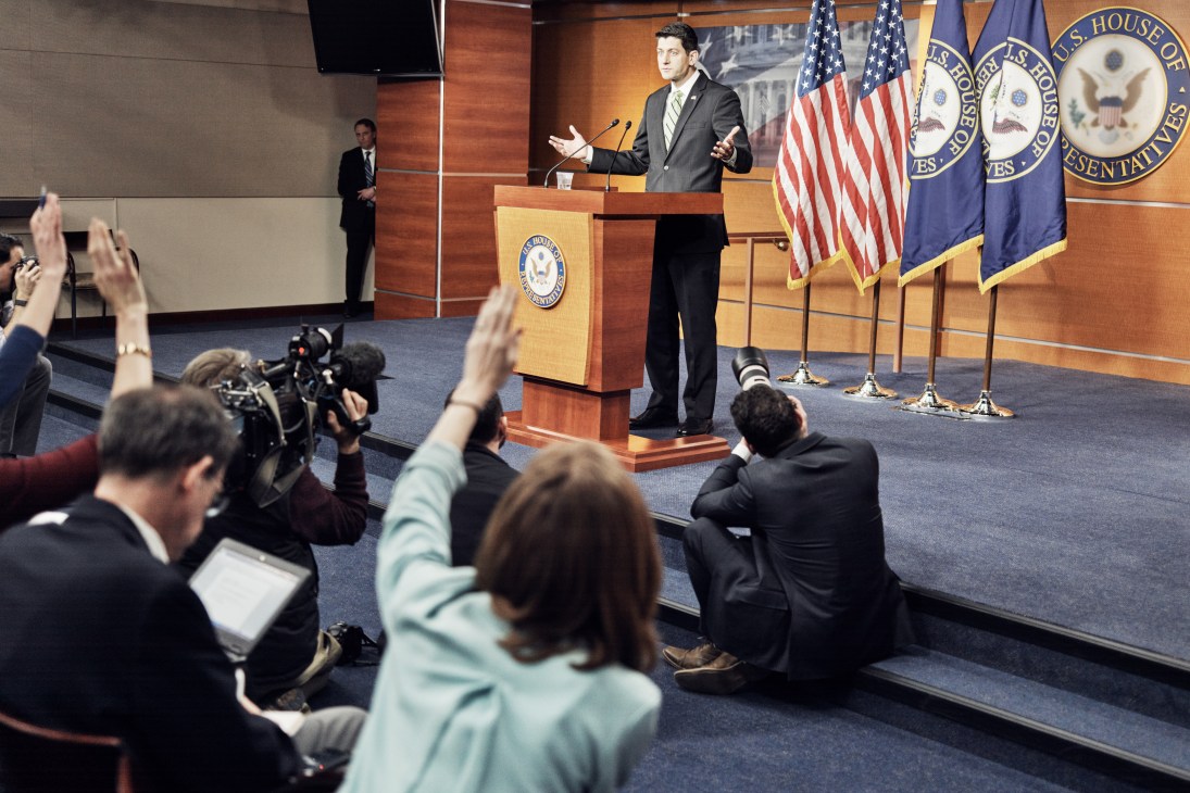 Journalist raises hand to ask question at US House press conference podium with American flags.