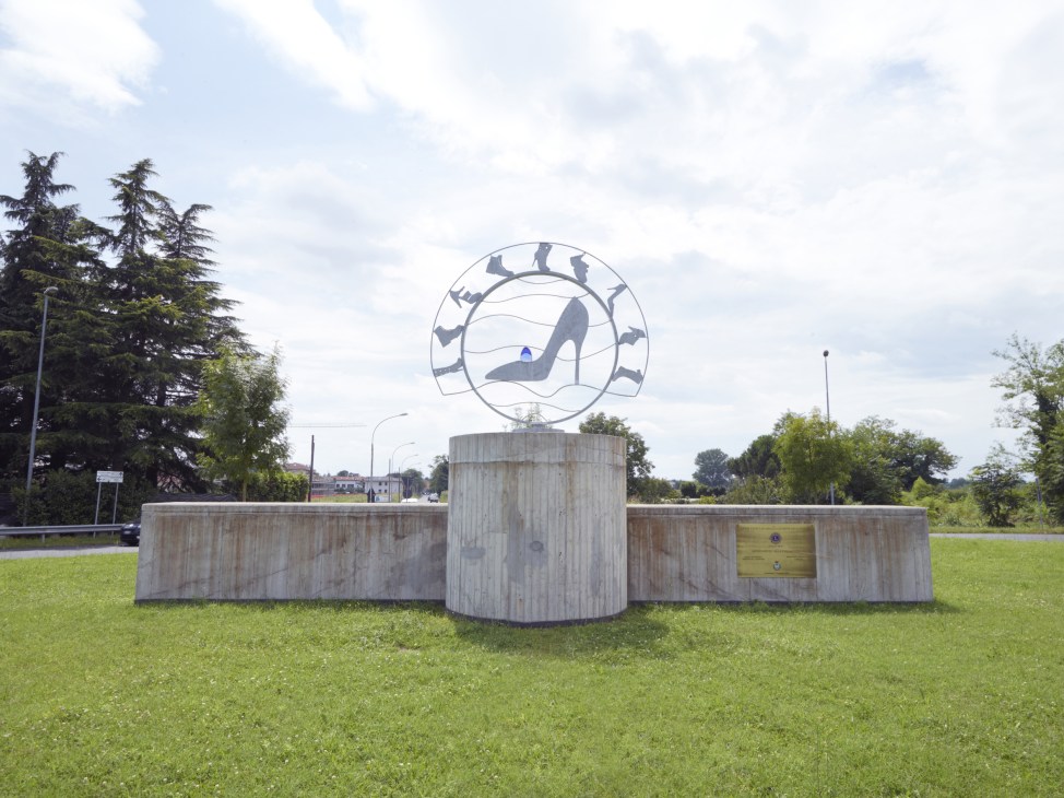 Monument to Parabiago's shoe district featuring a circular emblem with shoe silhouettes on a cylindrical concrete base in a green field.