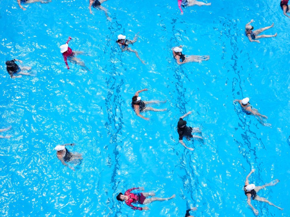 Multiple swimmers performing synchronized aqua-aerobics movements in a bright blue public pool during a group fitness class.