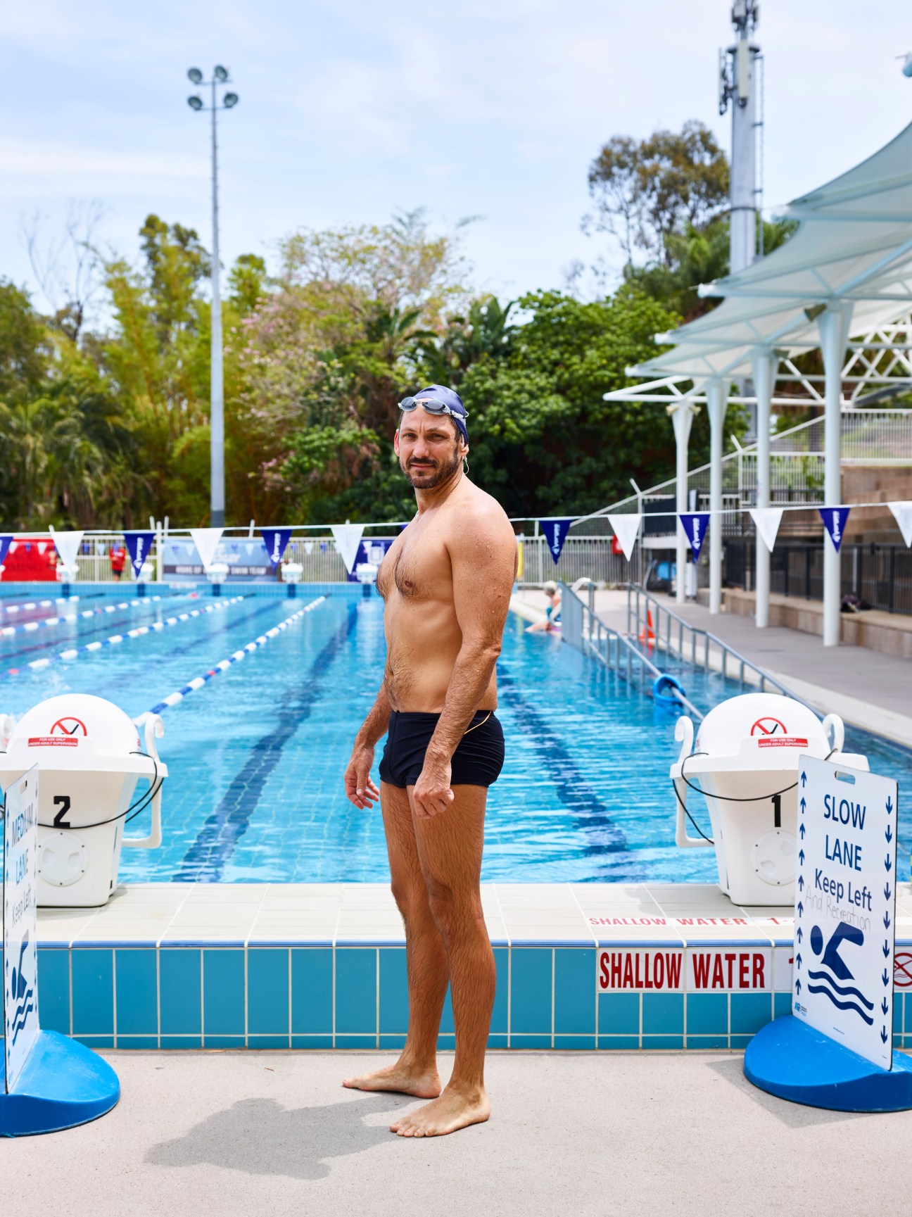 A male swimmer stands at the edge of a public swimming pool wearing goggles and black swim shorts.