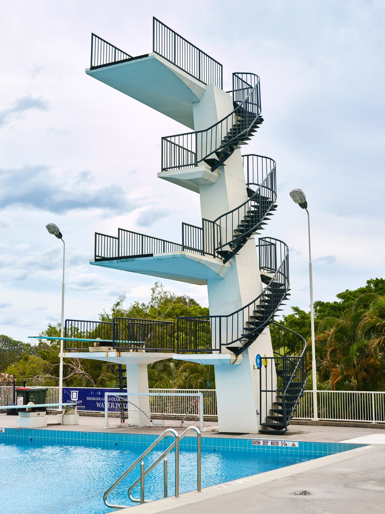 Tall white diving tower with blue platforms and black stairs next to a swimming pool.