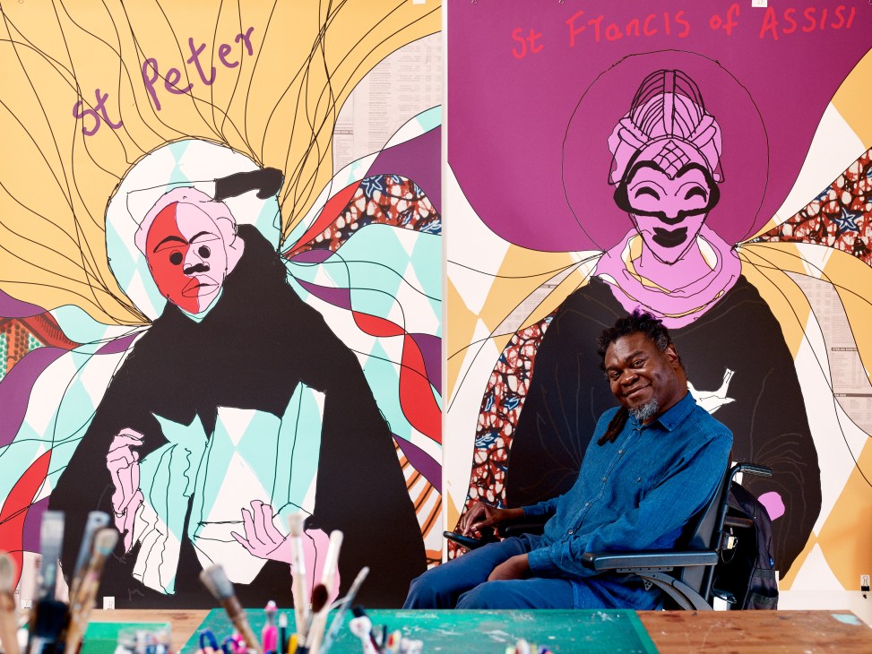 Yinka Shonibare sitting in front of colorful artwork blending African and Christian religious iconography with vibrant patterns and text.