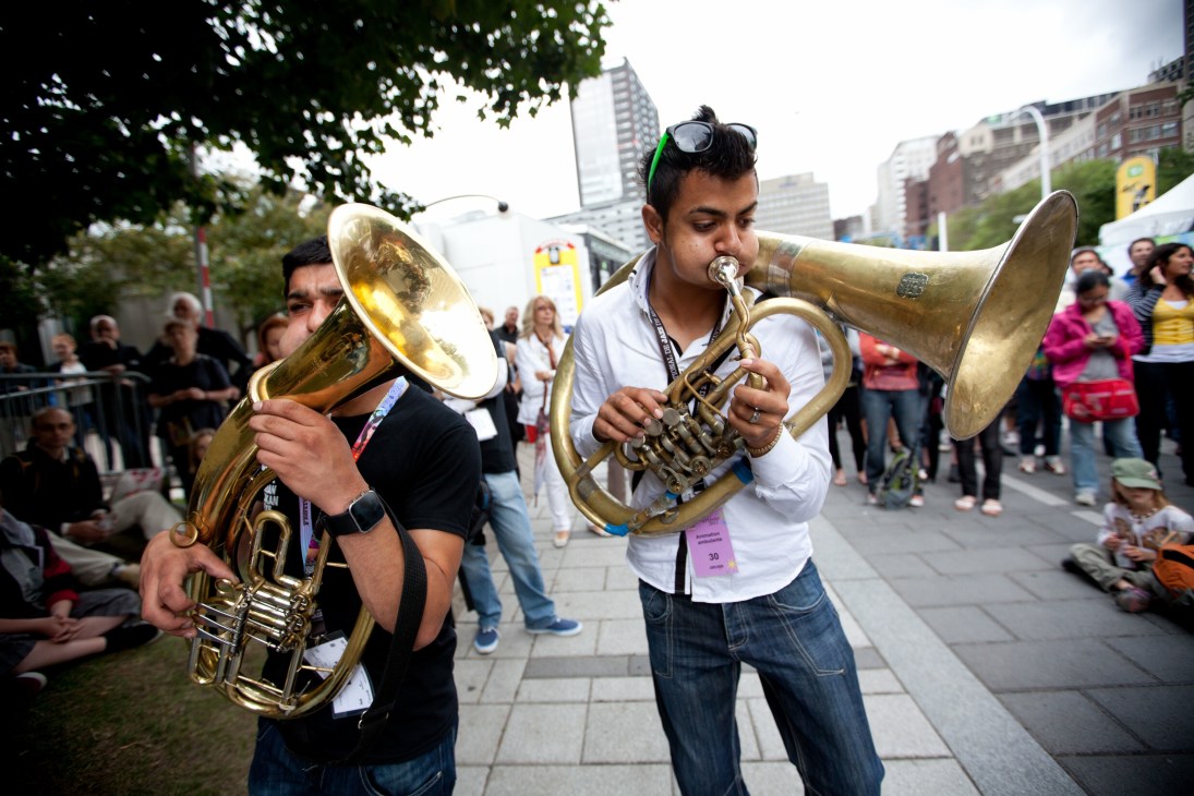 Two brass musicians performing with tuba and trumpet at an outdoor festival venue with city buildings in background.