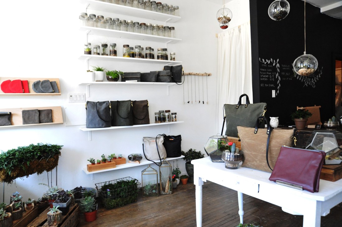 Crown Flora Studio storefront displaying waxed canvas bags on white shelves, potted plants, and hanging terrariums.