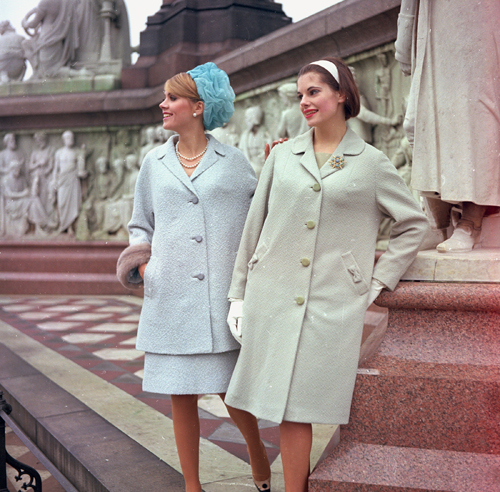 Two women in 1960s office attire wearing A-line coats, pillbox hats and kitten heels on classical steps.