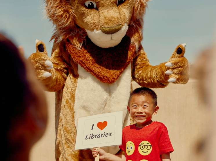 A lion mascot holds an "I love Libraries" sign while posing with a smiling young child in a red shirt.