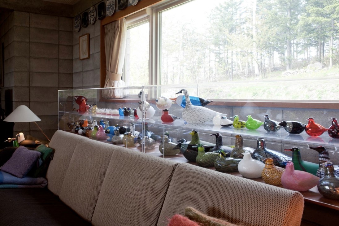 A light-filled room with a cream sofa in the foreground and glass display cases containing colorful glass bird sculptures and decorative objects.