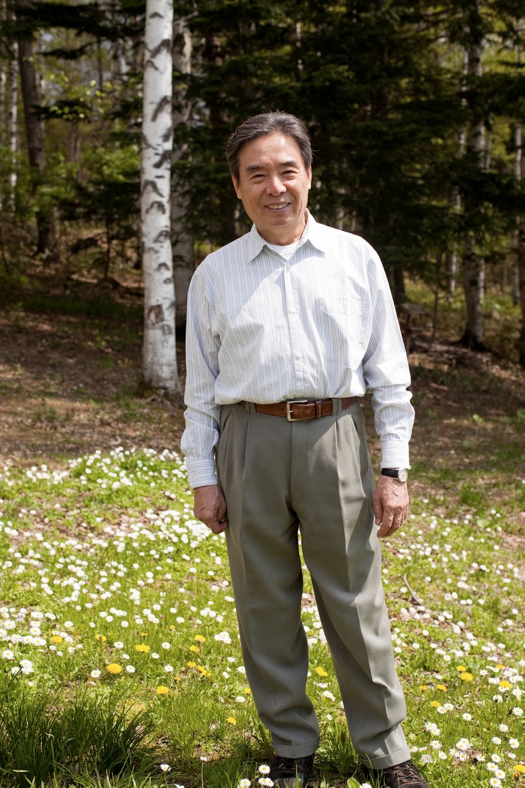 Noritsugu Oda stands smiling in a grassy field with white flowers and birch trees behind him.