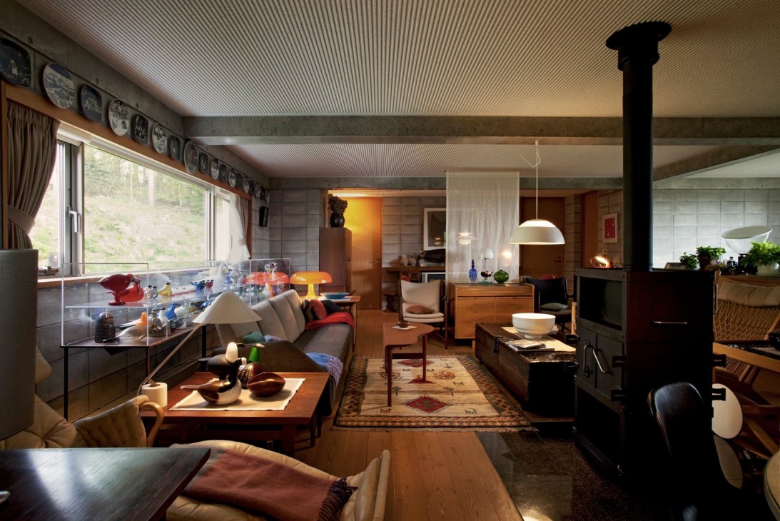 Second floor living room filled with Scandinavian mid-century furniture, pendant lighting, and decorative objects near large windows overlooking birch woods.