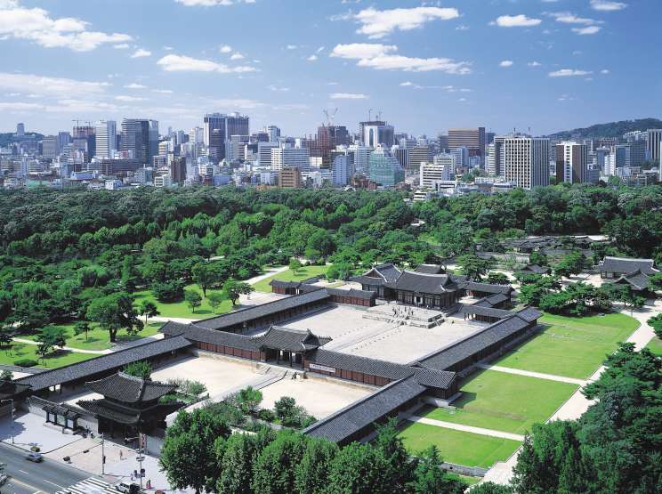 Changdeokgung Palace in Seoul with traditional Korean architecture surrounded by green gardens, overlooking modern city skyline.
