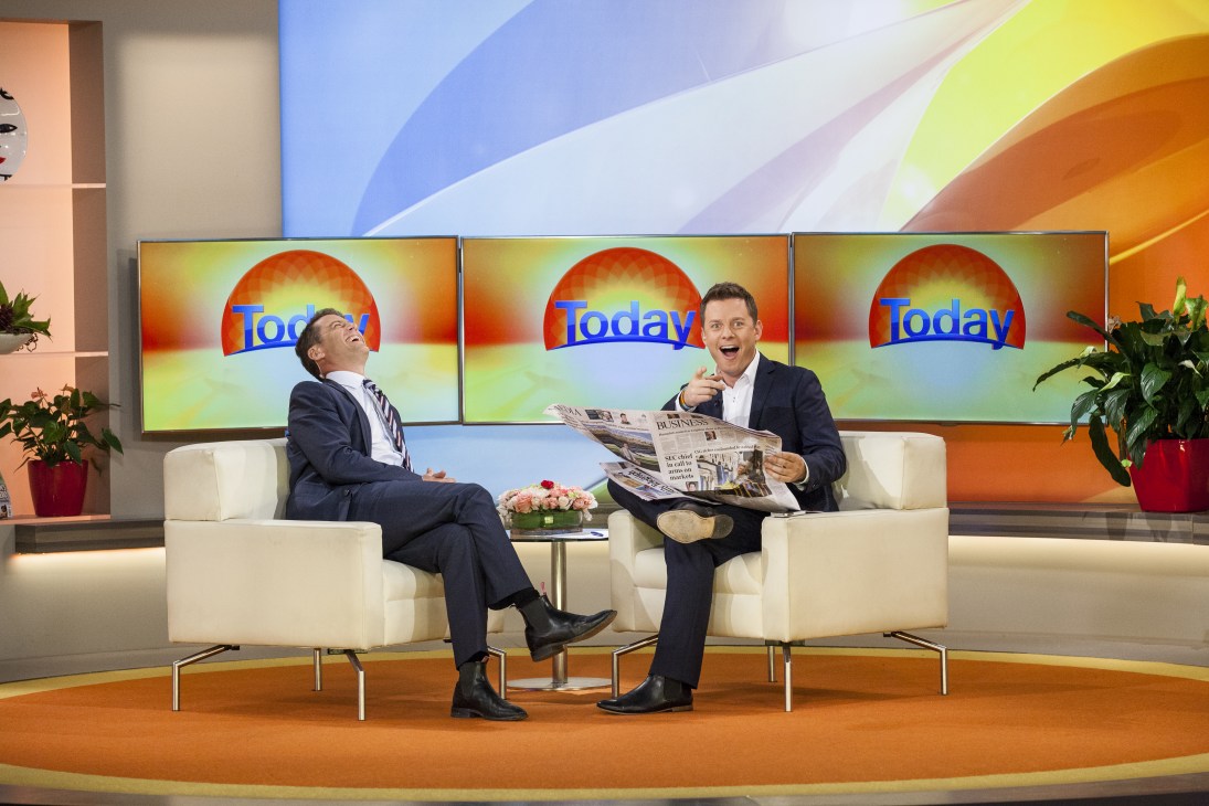Two men in suits sit on a white sofa on a TV studio set with 'Today' logos on screens behind them, one holding a newspaper.