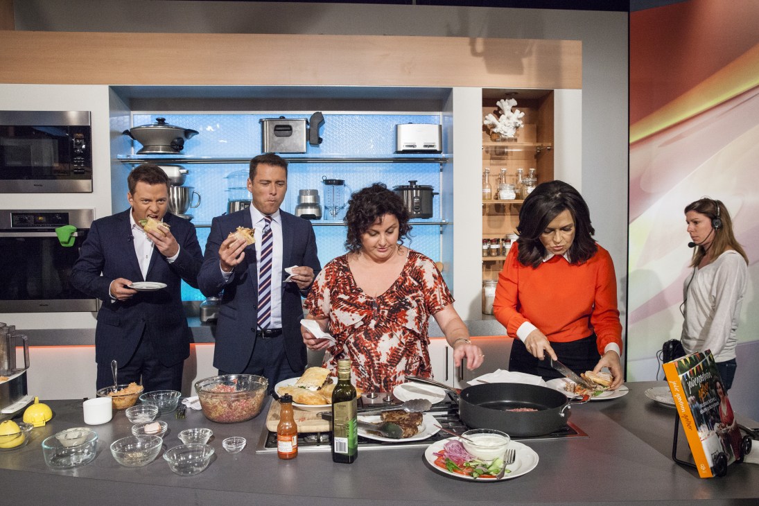 Five people stand around a kitchen counter on a TV set, with some eating from plates while others prepare food.