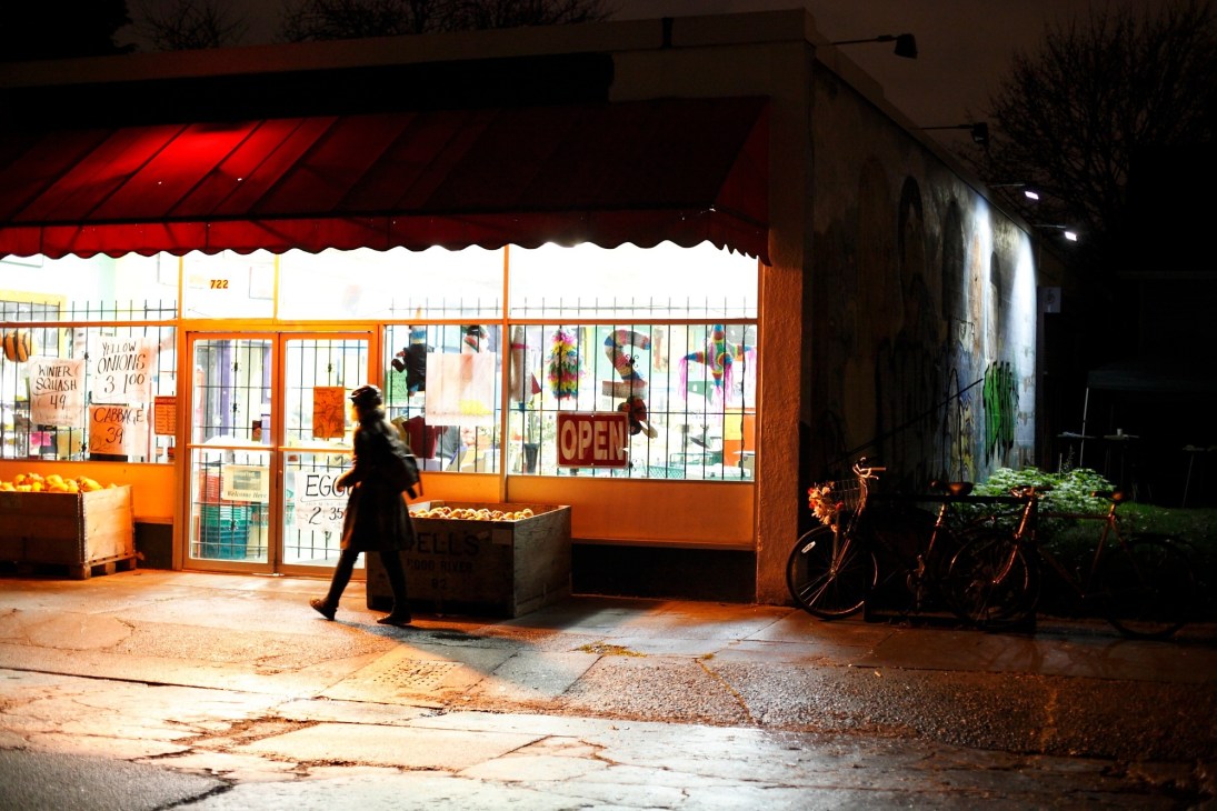 Illuminated storefront with red awning on a Portland street at night, person walking past.