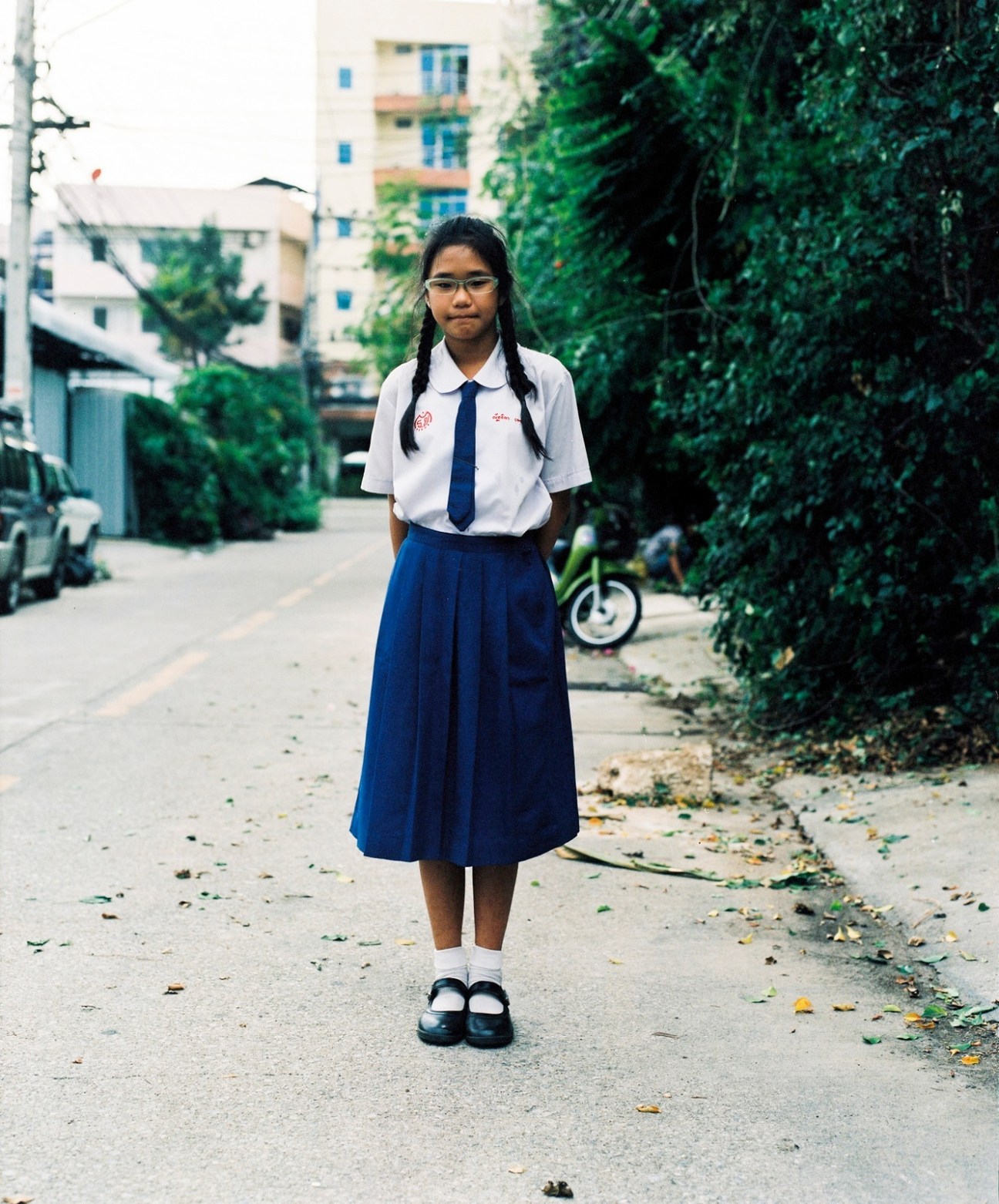 Thai schoolgirl in uniform standing on a residential street lined with ivy-covered walls.