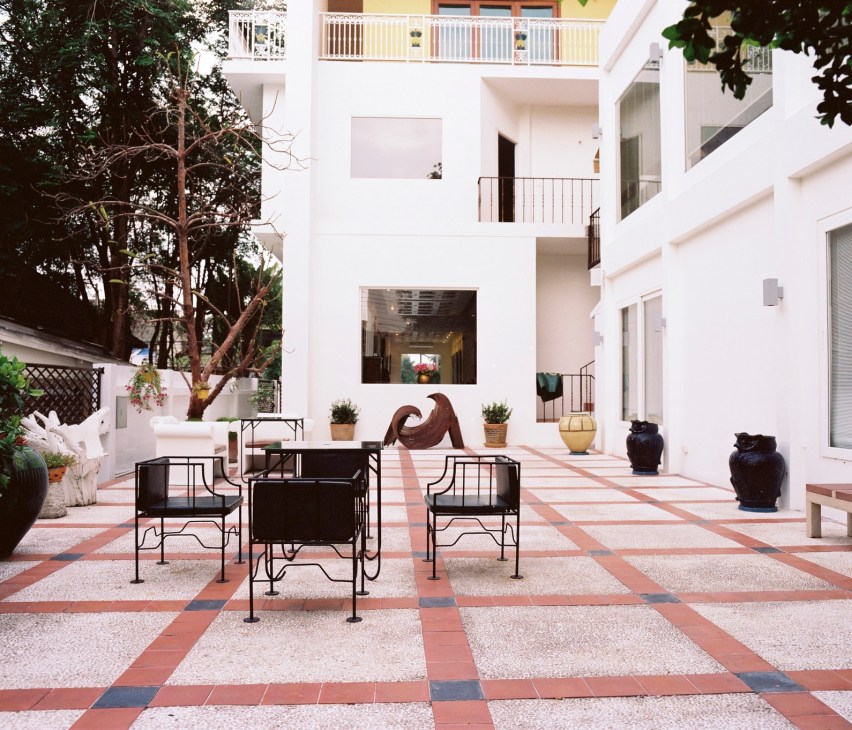 White modernist courtyard with geometric terracotta and blue floor tiles, black metal furniture, potted plants, and a sculptural wooden element.