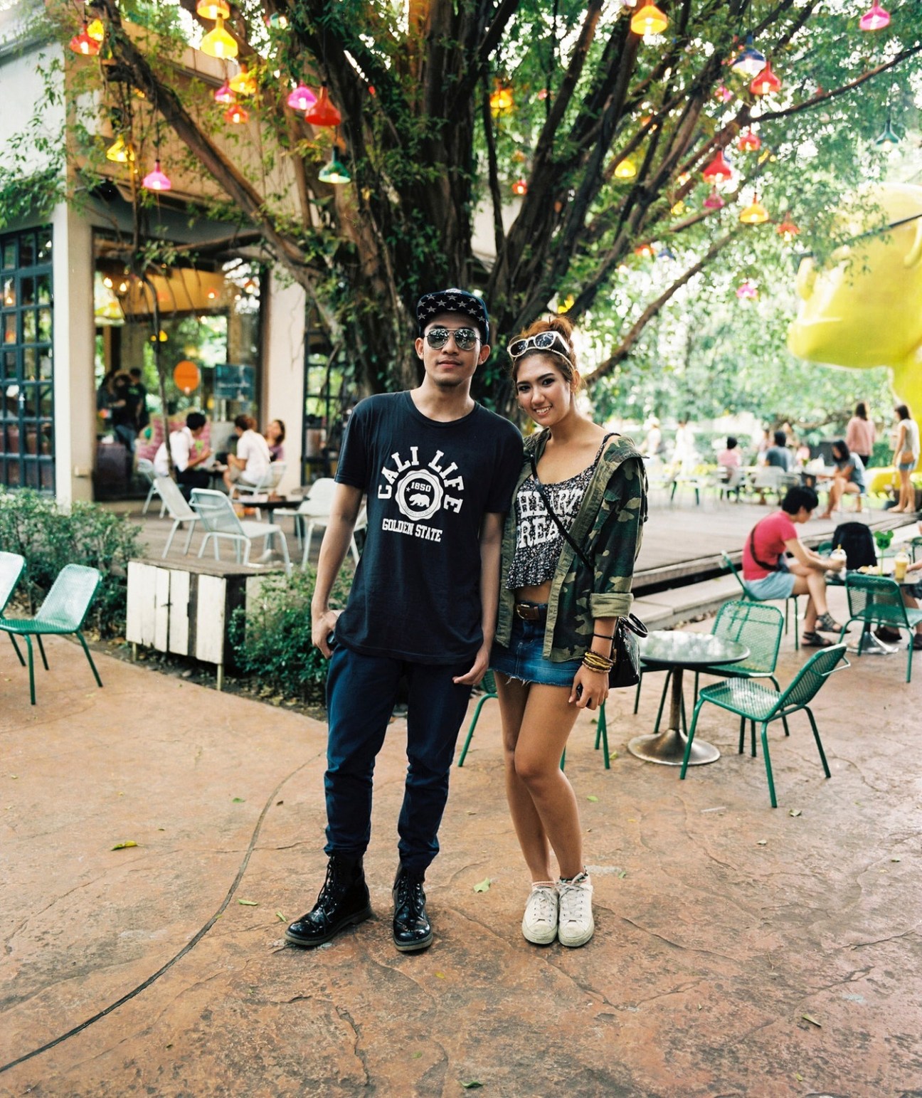 Young couple posing under a large tree with hanging lanterns in a public plaza with other visitors seated around them.