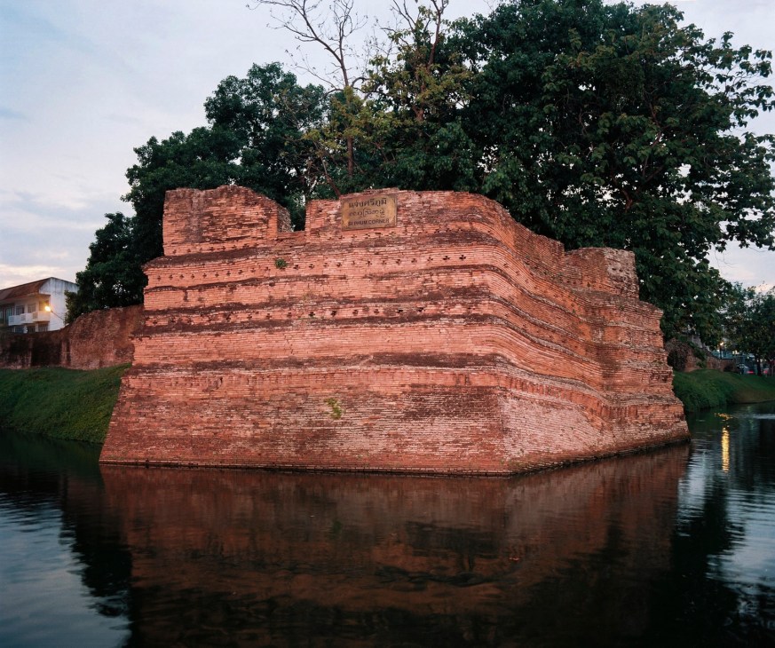 Red brick ruins of an ancient fortress structure surrounded by a moat with water, trees and buildings visible behind.